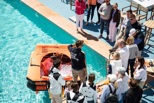 Sicherheitstraining mit einer aufgeblasenen orangefarbenen Rettungsinsel im Pool, Trainer demonstriert die Handhabung waehrend Teilnehmer vom Steg zuschauen