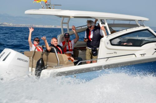 Froehliche Trainingsgruppe mit Schwimmwesten auf einem Motorboot auf dem Mittelmeer, Teilnehmer winken in die Kamera